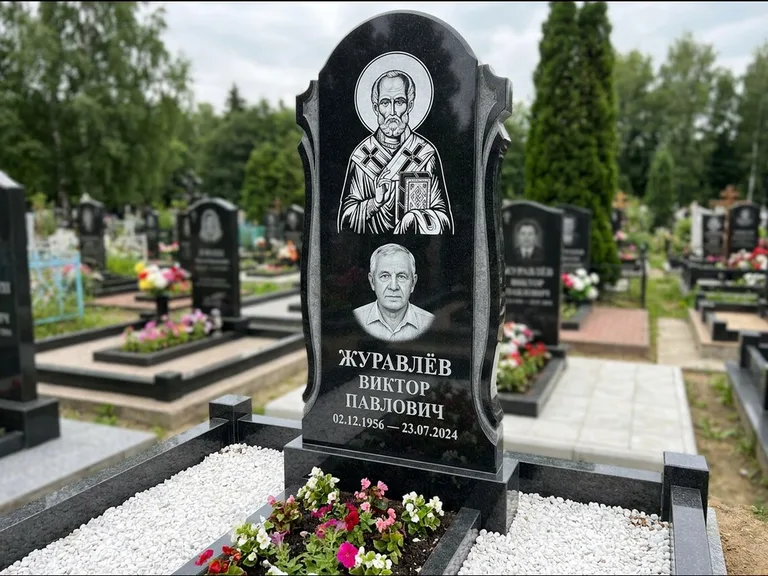 Памятник с гравировкой иконы: стела с образом в полный рост на обороте — гравировка образа Матроны Московской, низкие кусты спиреи в цвету, работа «Лебедева» в Сочи
